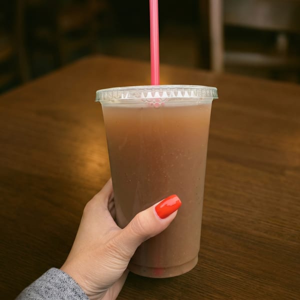 A hand with red nail polish holds a plastic cup with a brown drink and pink straw.