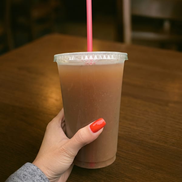 A hand with red nail polish holds a plastic cup with a brown drink and a pink straw.